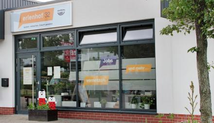 Das Bild zeigt die Front des Projekthauses erlenhof32. Zu sehen sind die Tür sowie die bis zum Boden reichende Fenster des Flachbaus. Auf der Fensterfront sind verschiedene Schriftzüge auf gelben und lila Grund zusehen. Über der Tür ist ein Schild mit der Aufschrift erlhof32 zu sehen. Vor dem flachen Gebäude steht ein Blumenkasten, weiter im Vordergrund rechts ein Baum und eine braune Holzbank auf dem sonst städtischen, gepflasterten Platz. Das Wetter ist grau. Das Projekthaus befindet sich am Schlaatzer Ma