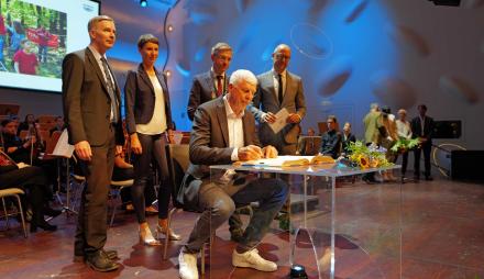 Der Geehrte Peter Schäperkötter sitzt an einem Tisch im Nikolaisaal und schreibt sich ins Goldene Buch der Stadt ein. Hinter ihm stehen Dr. Hagen Wegewitz, Mike Schubert und Sebastian Stielke.