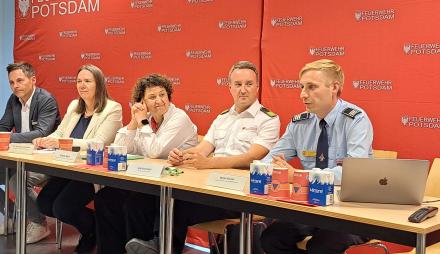 Fünf Menschen sitzen vor einer Pressewand der Feuerwehr Potsdam bei einer Pressekonferenz.