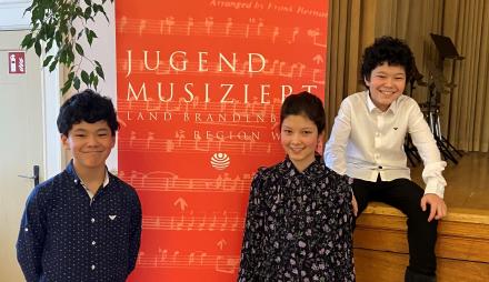 Die Drillinge Markus, Viola und Martin Xhyra vor dem Banner "Jugend musiziert"