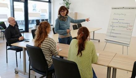 Eine Frau steht vor einem Flipchart und erklärt etwas. Ihr zugewandt sitzen drei Personen an Tischen, die ihr zuhören.