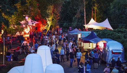 Ansicht des Localize Festivalgeländes in der Natur am Abend, mit bunter Beleuchtung und vielen Besuchenden