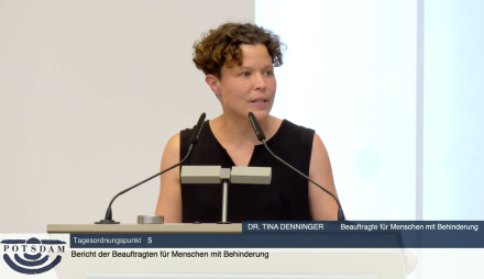 Das Bild zeigt Dr. Tina Denninger, Beauftragte für Menschen mit Behinderung der Landeshauptstadt Potsdam, bei einem Redebeitrag am Redepult während einer Sitzung der Stadtverordnetenversammlung (SVV). Sie spricht zum Tagesordnungspunkt 5 mit dem Titel „Bericht der Beauftragten für Menschen mit Behinderung“. Im Bild ist sie gestikulierend zu sehen, was auf ein engagiertes und erklärendes Sprechen hindeutet. Am unteren Bildrand ist das offizielle Potsdam-Logo eingeblendet.