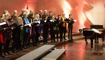 23 Mitglieder des Claudius-Ensembles singen auf der Bühne der Nikolaisaal; ein Flügel und ein Dirigierpult sind ebenfalls zu sehen.