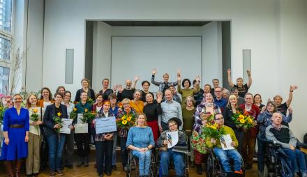 „Großes Gruppenfoto der Preisträgerinnen und Preisträger des Potsdamer Inklusionspreises 2025. Menschen mit und ohne Behinderung stehen und sitzen gemeinsam im Saal des Potsdam Museums, viele halten Blumen und Urkunden, alle lächeln und jubeln.“