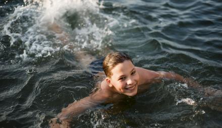 Eine Junge schwimmt in einem See und schaut lächelnd in die Kamera..