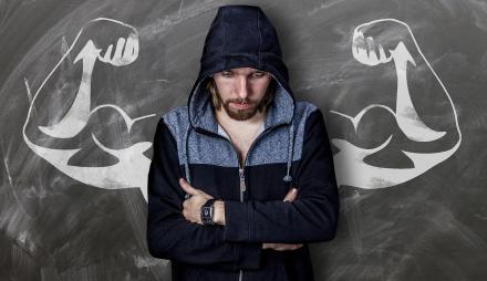 Ein Mann träumt davon, stark zu sein. Er ist niedergeschlagen, trägt einen Kapuzenpulli. Hinter ihm sieht man starke, muskelbepackte Arme, die ihm wie Flügel zu wachsen scheinen. Eine Kreidezeichnung auf grauem Grund im Bildhintergrund.
