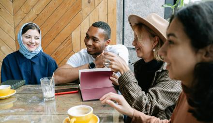 Vier Personen sitzen zusammen an einem Tisch. Sie lachen vor ihnen stehen Tassen und ein Glas. Eine Person schaut aiuf ein Tablet. Eine Frau trägt ein Kopftuch, auch ist ein Mann mit dunkler Hautfarbe dabei. Das Bild vermittelt Diversität und Gemeinschaft.