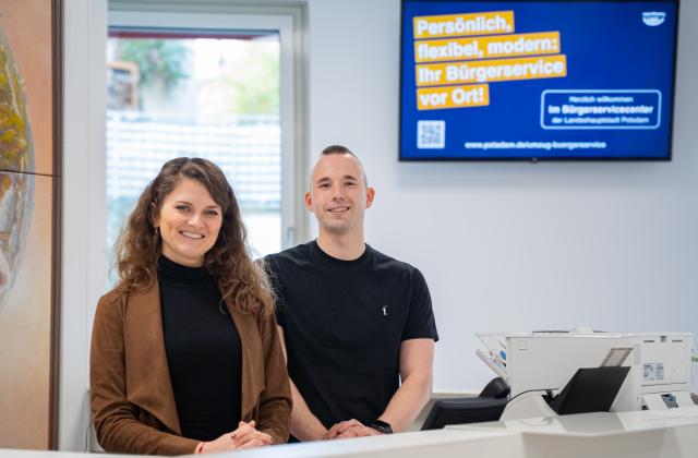 Zwei Mitarbeitende des Bürgerservice hinter einem Schalter schauen in die Kamera.