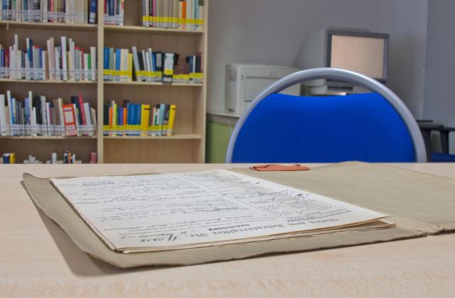 auf dem Tisch des Lesesaals im Stadtarchiv Potsdam liegt eine aufgeschlagene Akte, im Hintergrund sieht man einen Teil der Archivbibliothek und es leuchtet der Monitor eines Mikrofilmlesegerätes