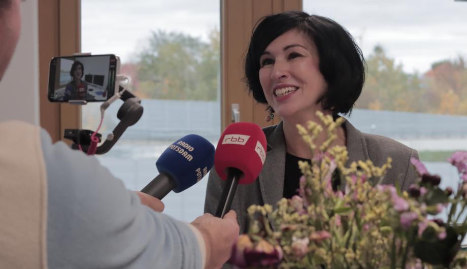 Noosha Aubel steht in ihrem Büro und gibt Interviews. Vor ihrem Mund sind Mikrofone des rbb und von Radio Potsdam. Im Hintergrund ist die Nuthestraße aus ihrem Bürofenster zu erkennen.