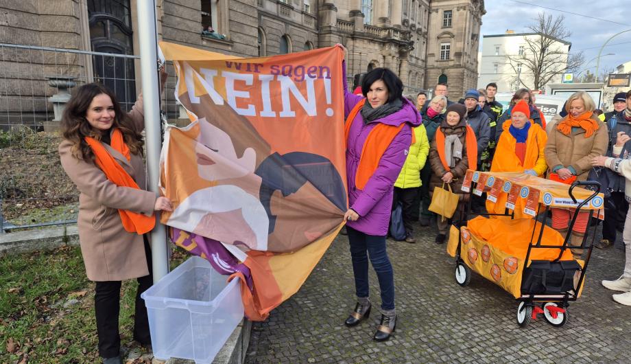 Eine Gruppe von Menschen steht vor dem historischen Rathausgebäude in Potsdam. Mehrere Personen tragen orangefarbene Schals und halten eine große Fahne mit der Aufschrift „Wir sagen NEIN!“ hoch. Die Szene zeigt die Hissung der UN-Women-Fahne zum Internationalen Tag gegen Gewalt an Frauen. Im Hintergrund sind die Fassade des Gebäudes und der grüne Turm des Rathauses zu sehen.