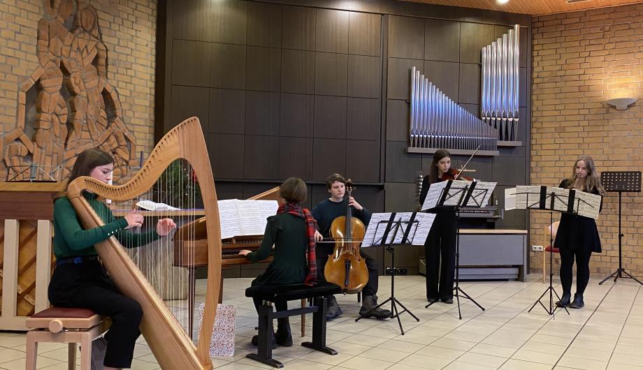 In einer etwas nüchternen Kirche musiziert ein kleines Barock-Ensemble mit jungen Spielerinnen und Spielern. Es werden eine Harfe, ein Cembalo, ein Cello, Violine und Flöte verwendet.