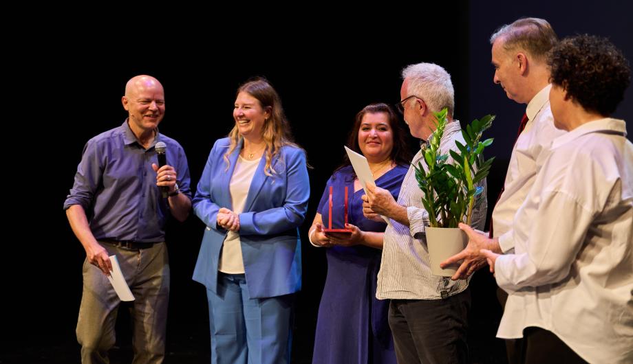 Das Foto zeigt eine fröhliche Gruppe von sechs Personen auf einer Bühne während einer Preisverleihung. Eine Person spricht ins Mikrofon, die anderen lächeln und halten Urkunden, Blumen und eine Trophäe in den Händen. Die Stimmung ist herzlich und feierlich – der Moment wirkt wie die Übergabe oder Würdigung eines Preises.