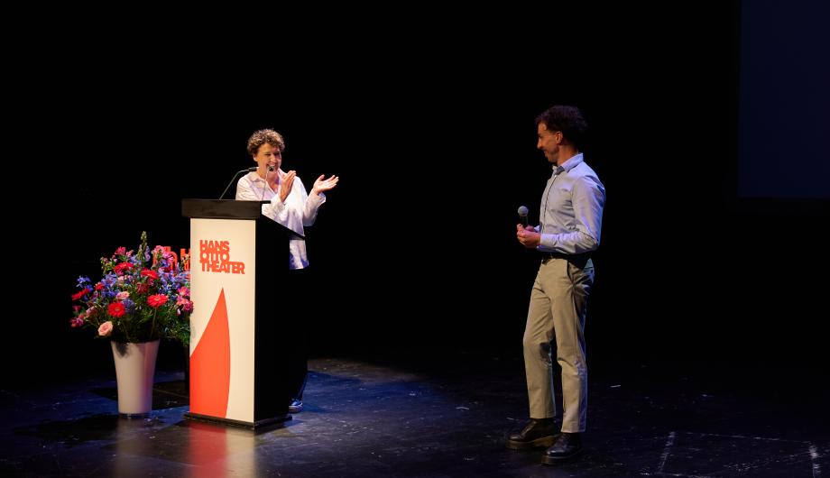 Das Foto zeigt eine Person am Rednerpult auf einer Bühne des Hans Otto Theaters, die in Richtung eines Mannes spricht und gestikuliert. Neben dem Pult steht ein großer Blumenstrauß. Die Szene wirkt lebendig und herzlich – offenbar handelt es sich um einen Moment während einer Veranstaltung oder Preisverleihung.