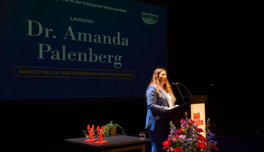Das Foto zeigt eine Frau am Rednerpult auf der Bühne des Hans Otto Theaters. Hinter ihr ist eine große Leinwand mit der Aufschrift „Laudatio: Dr. Amanda Palenberg, Beauftragte für Migration und Integration“ zu sehen. Auf einem Tisch neben dem Pult stehen rote Trophäen und Pflanzen, vor dem Podium ein Blumengesteck. Die Szene zeigt einen Moment während der feierlichen Verleihung des Potsdamer Integrationspreises.