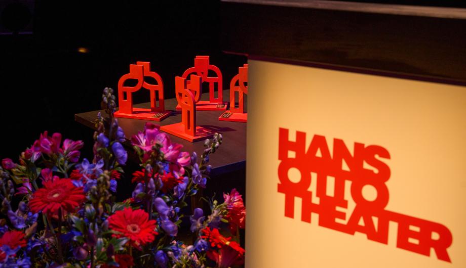 Das Foto zeigt mehrere rote Trophäen des Potsdamer Integrationspreises, die auf einem Tisch im Hans Otto Theater arrangiert sind. Im Vordergrund sind farbenfrohe Blumen zu sehen, rechts der leuchtende Schriftzug „Hans Otto Theater“ auf einem Podium. Das Bild fängt die festliche Atmosphäre der Preisverleihung ein.