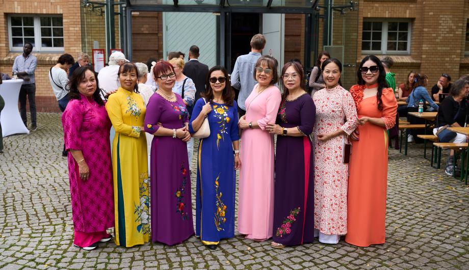 Das Foto zeigt eine Gruppe von acht Frauen, die in farbenfrohen, traditionellen Kleidern zusammenstehen und lächeln. Sie posieren im Freien auf einem gepflasterten Hof, im Hintergrund sind weitere Gäste und Sitzbänke zu sehen. Die Szene vermittelt festliche Stimmung und kulturelle Vielfalt – ein Moment der Begegnung im Rahmen der Potsdamer Integrationspreisverleihung.
