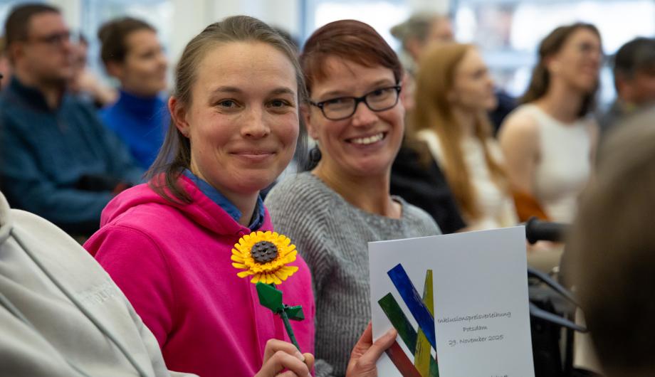 „Zwei lächelnde Frauen sitzen im Publikum. Eine von ihnen hält eine Sonnenblume und eine Urkunde der Inklusionspreisverleihung in der Hand. Im Hintergrund sitzen weitere Gäste des Festakts.“