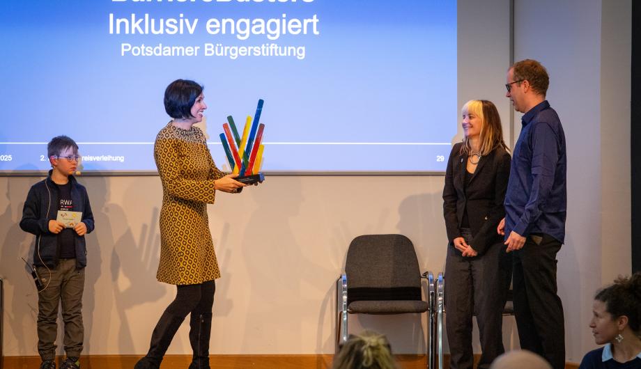 „Bei der Preisverleihung überreicht Oberbürgermeisterin Noosha Aubel den BarriereBusters die bunte Inklusionspreis-Skulptur. Zwei Vertreter der BarriereBusters stehen lächelnd auf der Bühne, während ein Kind daneben zuschaut. Im Hintergrund ist die Folie mit dem Projektnamen eingeblendet.