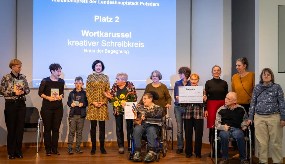 Gruppenfoto zur Verleihung des 2. Platzes beim Inklusionspreis Potsdam: Das Wortkarussell – kreativer Schreibkreis – steht gemeinsam mit Oberbürgermeisterin Noosha Aubel, Moderatorin und weiteren Beteiligten auf der Bühne. Eine Person im Rollstuhl hält die Urkunde, eine weitere Person ein großes Preisgeld-Schild. Im Hintergrund zeigt die Leinwand die Auszeichnung ‚Platz 2 – Wortkarussell‘.
