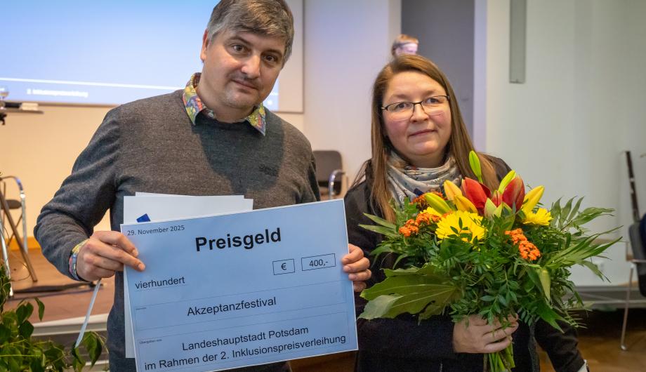 Zwei Vertreter*innen des Akzeptanzfestivals stehen nebeneinander und halten den Sonderpreis des Inklusionspreises Potsdam: einen großen symbolischen Scheck über 400 Euro sowie einen Blumenstrauß. Beide lächeln in die Kamera.