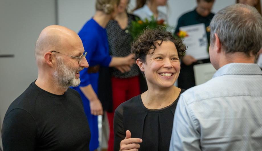 Drei Personen der Jury, Tina Denninger, Dirk Tannert und Tobias Straub,  stehen im Gespräch zusammen und lachen. Im Hintergrund sind weitere Gäste der Preisverleihung zu sehen, einige halten Blumen und Urkunden