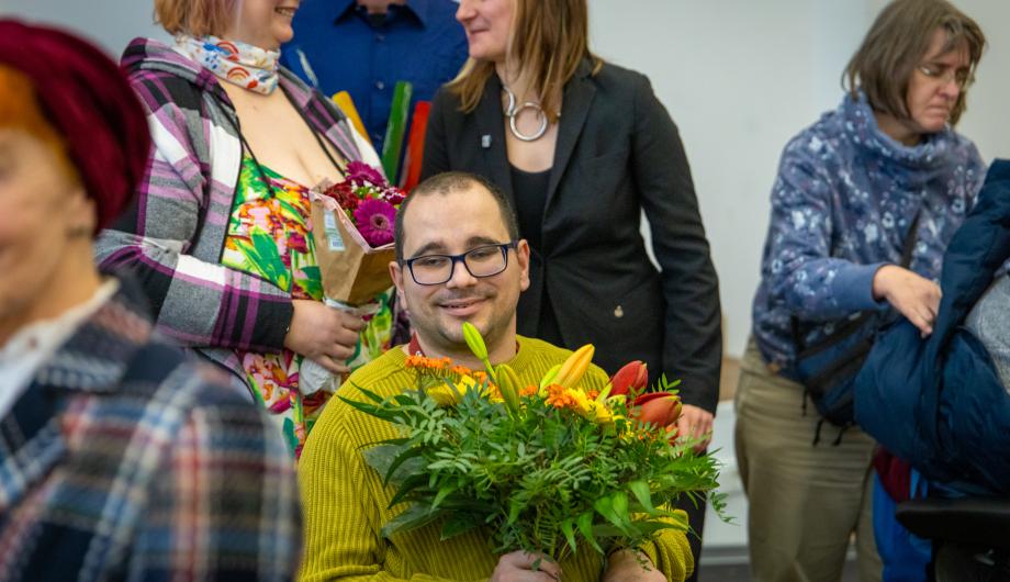 Jan Krech vom Stammtisch Inklusiv, Gewinner des 3. Platzes, sitzt mit einem bunten Blumenstrauß lächelnd im Vordergrund. Hinter ihm stehen weitere Teilnehmende der Preisverleihung mit Blumen und Urkunden.