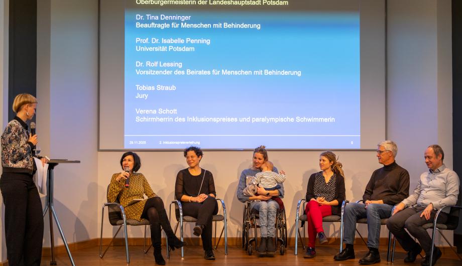 Podiumsdiskussion beim 2. Potsdamer Inklusionspreis: Sechs Personen sitzen auf der Bühne, darunter Oberbürgermeisterin Noosha Aubel, Dr. Tina Denninger, Prof. Dr. Isabelle Penning, Dr. Rolf Lessing, Tobias Straub und Verena Schott. Links steht die Moderatorin mit Mikrofon. Hinter ihnen ist eine große Leinwand mit den Namen der Podiumsteilnehmenden eingeblendet.