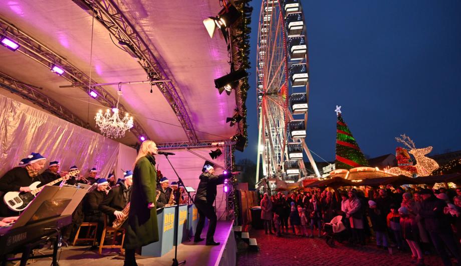 Ein Bild mit spektakulärer Weite zeigt den Potsdamer Weihnachtsmarkt: Links die beleuchtete Bühne mit einer Bigband, rechts viele Menschen auf dem Weihnachtsmarkt in blauer Stunde, und in der Mitte leuchtet das große Riesenrad.