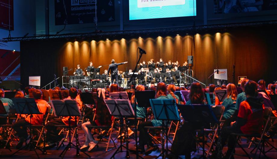 Kinder aus Musikklassen lauschen dem LAJJAZZO Junior Im Vordergrund haben sich viele Kinder von der Kamera weggedreht. Denn hinter Ihnen steht die große Jugendbigband Lajjazzo Junio und spielt.