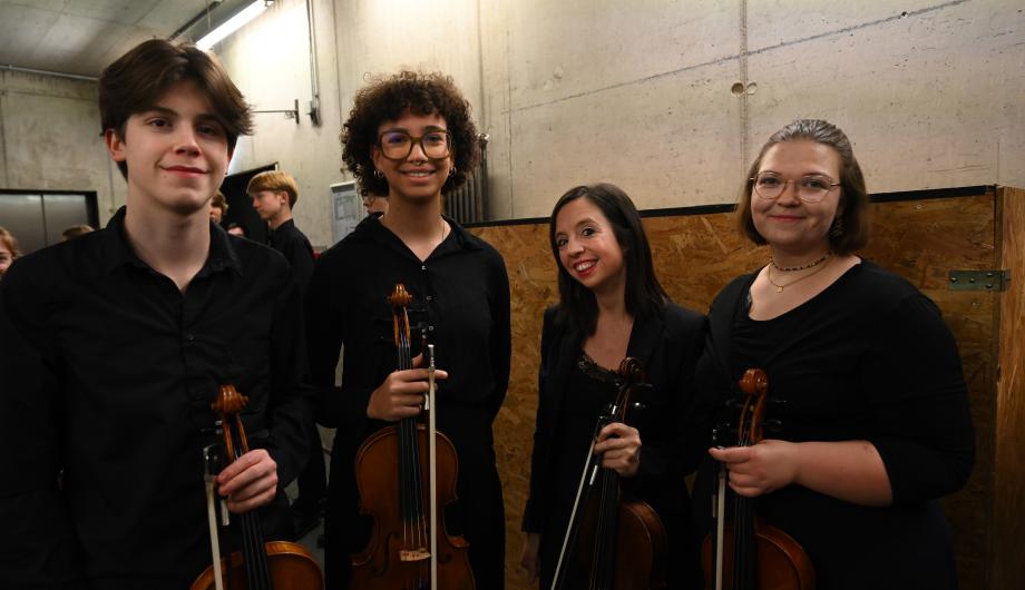 Ohne Bratschen geht es nicht! Vier junge Menschen halten eine Bratsche in ihren Händen. Sie lächeln in die Kamera und freuen sich auf ihr Konzert