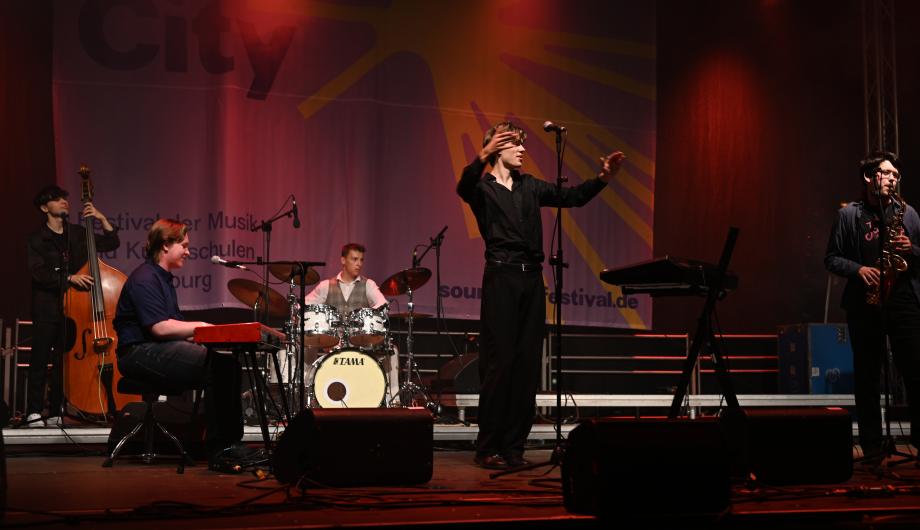 Eine Pop-Jazzband mit 5 jungen Männern performed auf professionell beleuchteter Bühne - der Sänger fordert zum Mitklatschen auf