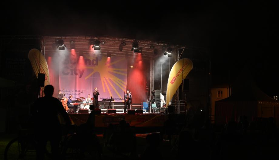 Eine Pop-Jazzband mit 5 jungen Männern performed auf einer weiter entfernten - professionell beleuchteter Bühne - der Sänger fordert zum Mitklatschen auf
