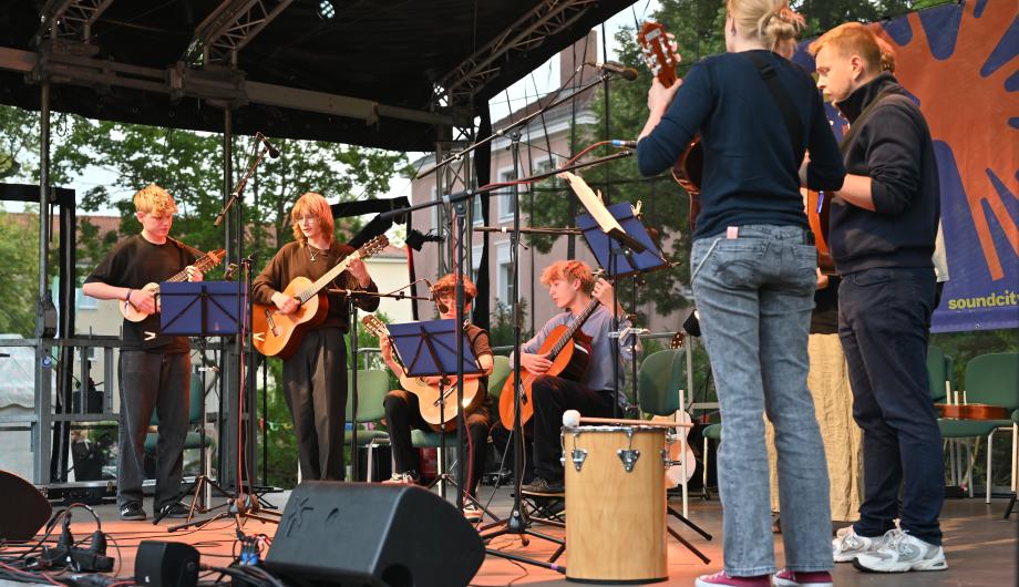 Ein 8-köpfiges altersgemischtes Gitarrenensemble spielt auf einer abendlich beleuchteten Open Air Bühne