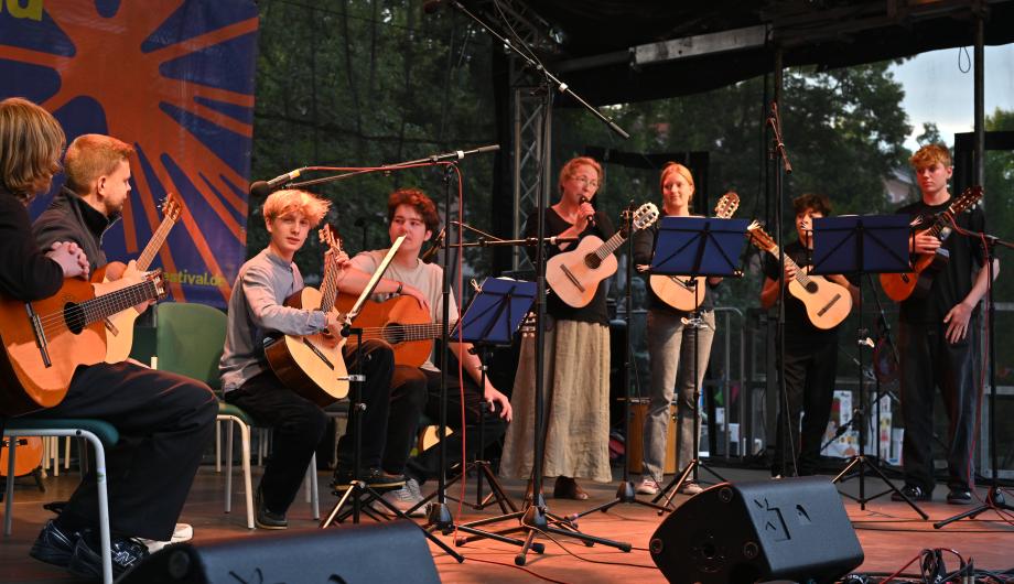 Ein 8-köpfiges altersgemischtes Gitarrenensemble spielt auf einer abendlich beleuchteten Open Air Bühne - die Leiterin macht eine Ansage