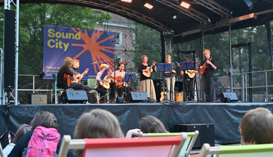 Ein 8-köpfiges altersgemischtes Gitarrenensemble spielt auf einer abendlich beleuchteten Open Air Bühne - davor sitzt Publikum im LIegestuhl
