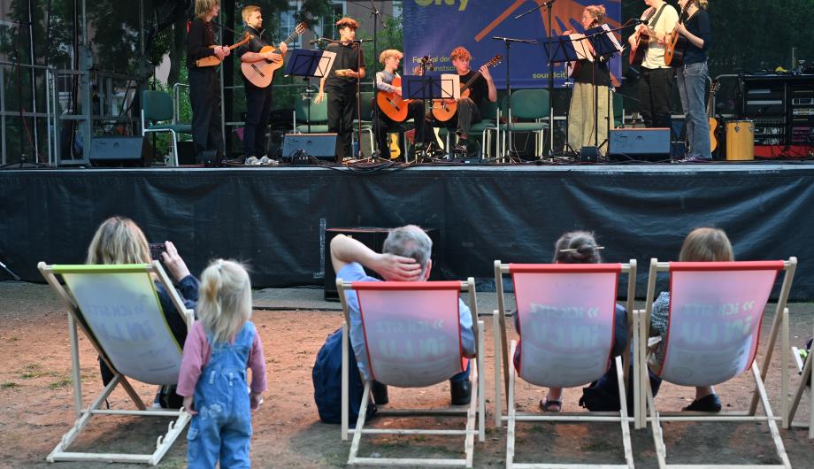 Ein 8-köpfiges altersgemischtes Gitarrenensemble spielt auf einer abendlich beleuchteten Open Air Bühne - davor sitzt Publikum im LIegestuhl - ein Kind bleibt stehen und hört zu