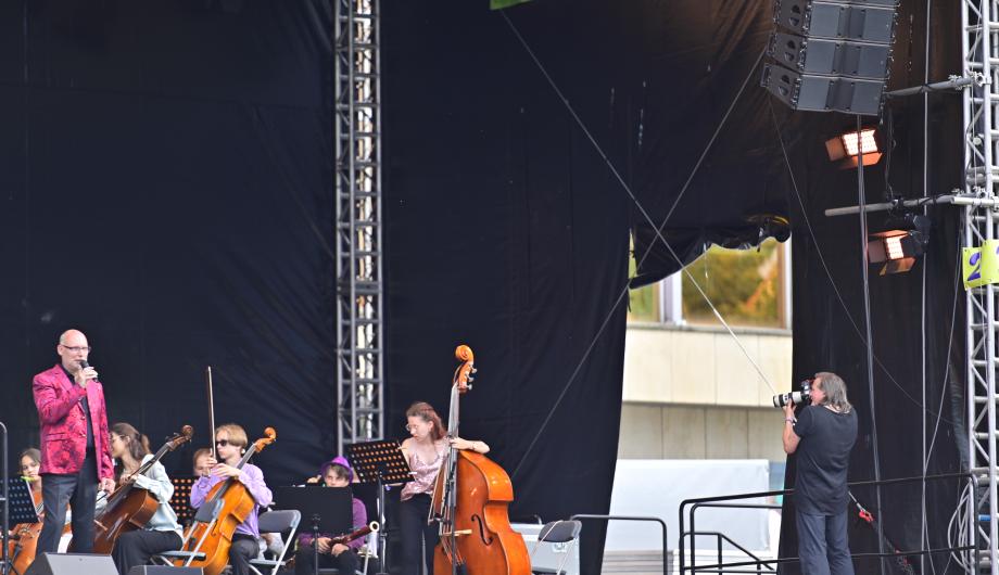 Auf einer Open Air-bühne steht ein Moderator vor einem Orchester. Rechts im Bild steht der Fotograf des Verbands der Musik- und Kunstschulen der hier bei seiner Arbeit gezeigt werden soll