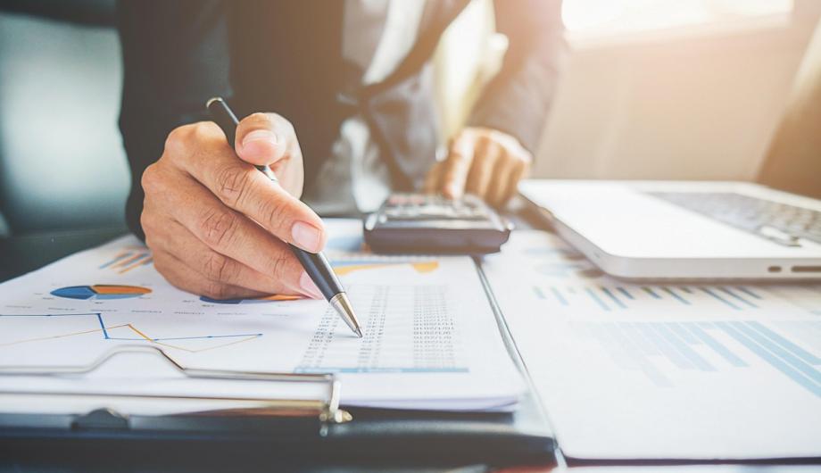 Hand eines Menschen am Schreibtisch mit Unterlagen, Laptop und Taschenrechner