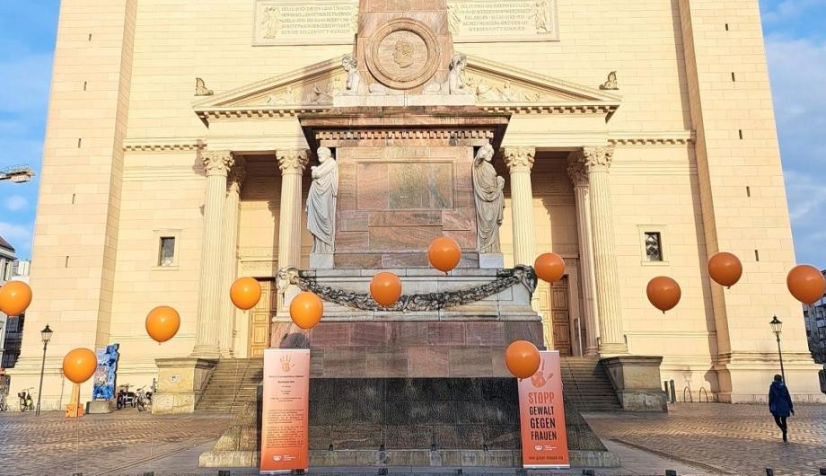 Auf dem Alten Markt in Potsdam stehen vor der Nikolaikirche mehrere orangefarbene Ballons als Teil einer Installation zum Internationalen Tag gegen Gewalt an Frauen. Im Vordergrund sind zwei Infotafeln aufgestellt.