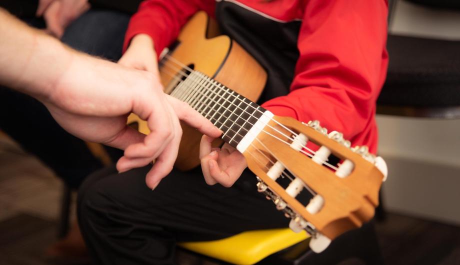 Unterrichtssituation mit einer akustischen Gitarre. Lehrkraft und Spieler sind nicht zu erkennen. Eine Hand weist auf eine Stelle auf dem Gitarrengriffbrett.