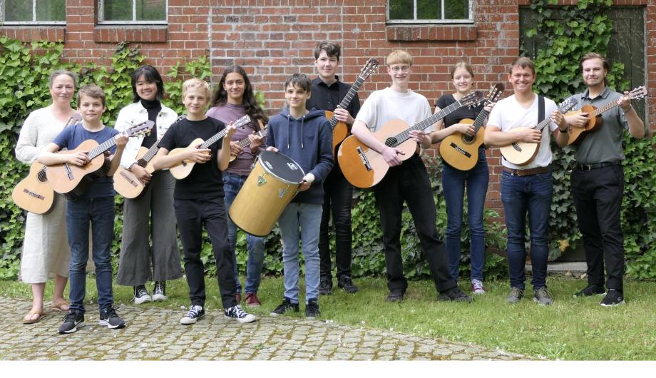 Les Bouffons - das folkloristische Gitarrenensemble Die 11 Musikerinnen und Musiker eines folkloristischen Gitarrensembles stehen auf Rasen vor der Backsteinmauer im Hinterhof der Musikschule Potsdam