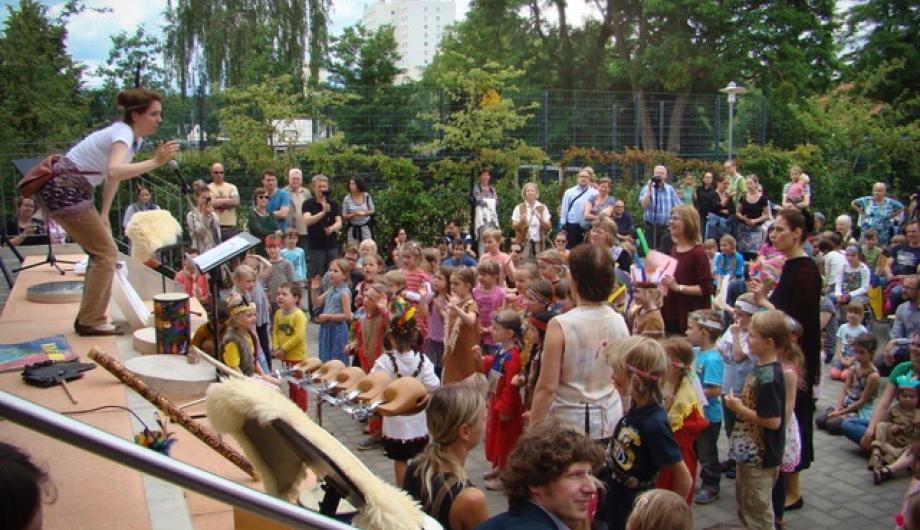 Mitmachaktion auf dem Schulhof - Standort Galileistraße Blick auf einen Schulhof mit grüner Bepflanzung im Hintergrund. Kinder und Eltern schauen auf eine Bühne, von der eine musikalische Mitmachaktion angeleitet wird.