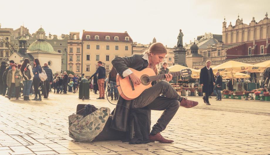 In romanitischem Sepia getöntes Bild. Ein etwas altmodisch gekleideter junger Mann sitzt auf einem alten Marktplatz und spielt seine Gitarre.