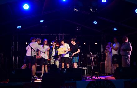 Die Rockband Solid Preachers Club steht glücklich nach ihrem Auftritt auf einer bunt beleuchteten Bühne. Ein Moderator überreicht einen Sonderpreis, die Jury applaudiert.