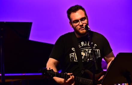 Ein Junger Mann spielt E-Gitarre und singt vor lila leuchtendem Bühnenhintergrund