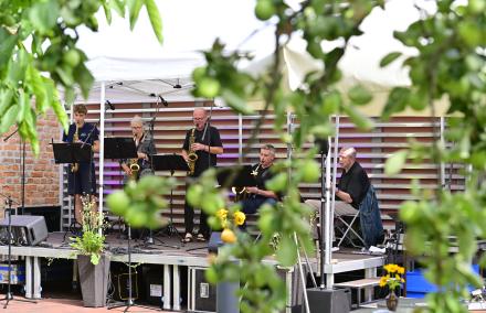 Durch das Grün von Bäumen ist eine Jazzcombo mit 4 Saxophonspielenden und einem E-Bassisten zu sehen. Die Gruppe ist sehr altersgemischt.