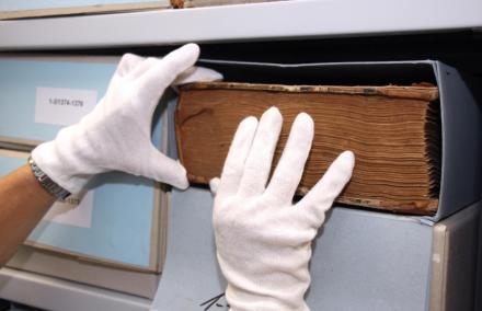 eine gebundene Archivale wird mit weißen Handschuhen aus dem Archivkarton genommen