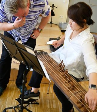 Die Spielweise der koreanischen Zither Gayageum Eine koreanische Frau erklärt die Spielweise ihres Saiteninstrumentes. Ein Komponist schaut neugierig
